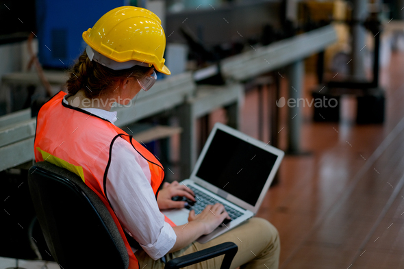 Pretty technician or engineer or worker sit on chair and use laptop in factory.