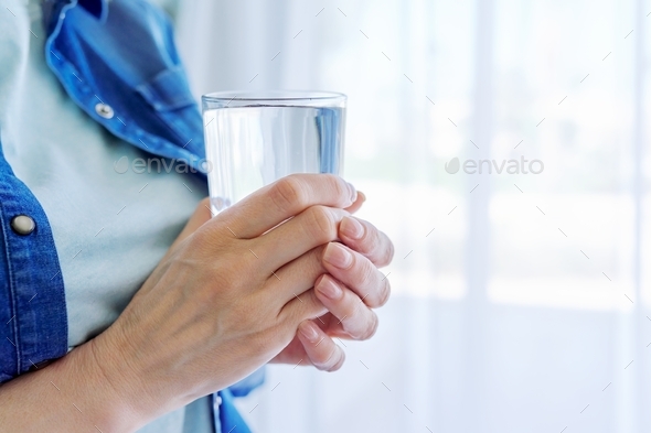 Close-up of middle-aged woman\'s hands holding glass of water, at home near window, copy space