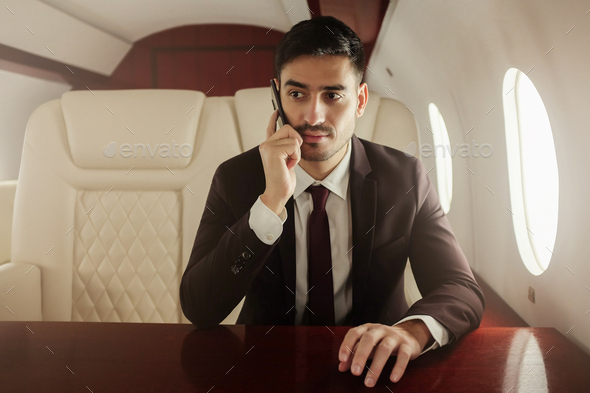 Rich business man sitting in private jet next to window, answering phone call
