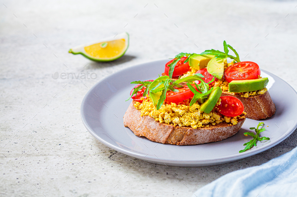 Tofu scrambled sandwich with avocado, arugula and tomato on gray plate. Healthy vegan food concept.