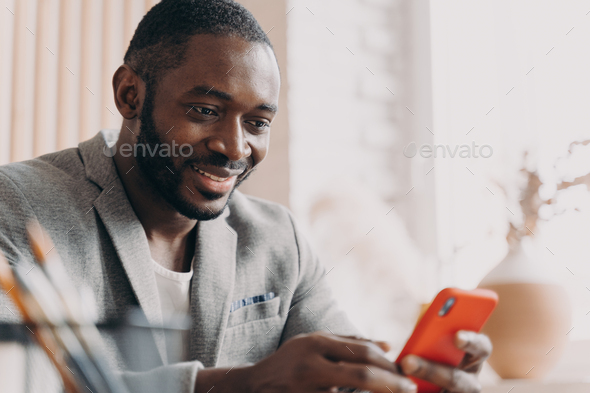 Smiling african american businessman using mobile, chatting online while sitting at office desk - Stock Photo - Images
