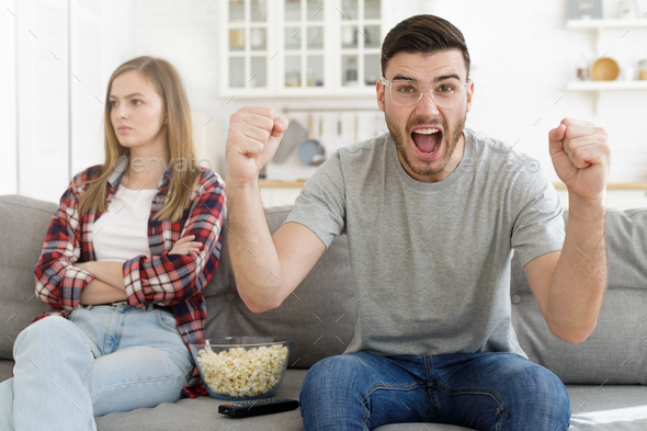 Young man watches sports game while his girlfriend sitting nearby is disappointed