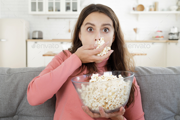 Young beautiful woman eats popcorn while watching TV completely absorbed by series