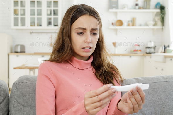 Sad young pregnant woman sits on couch at home checking pregnancy test results