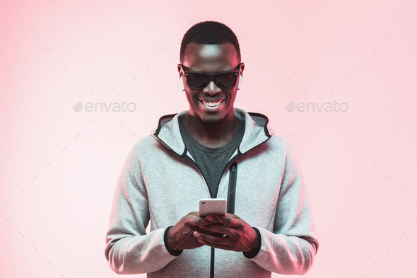 Portrait of African guy listening to music via wireless earbuds and using smartphone