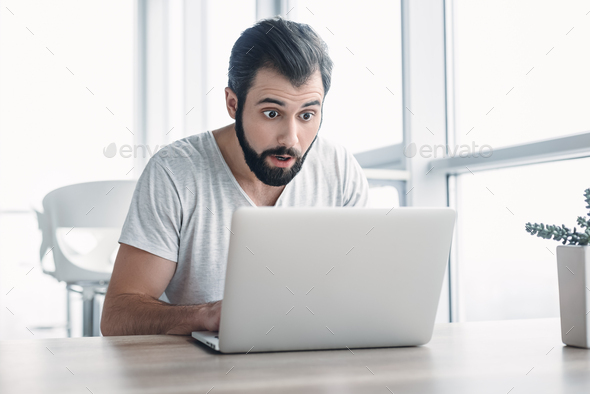 Young stressed businessman sitting with laptop with shocked facial expression. Surprised business