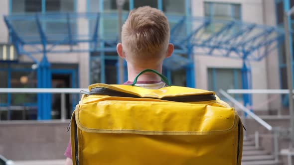 Young Man in Covid19 Face Mask with Yellow Food Delivery Bag Turning to Camera Standing in Front of alt