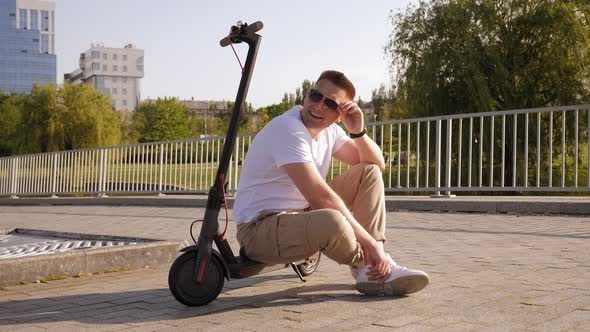 Fashionable Young Man in Sunglasses Is Sitting on an Electric Scooter in a Park. alt