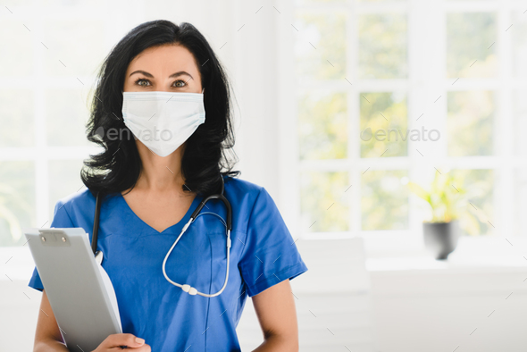 Caregiver in blue uniform face mask against coronavirus Covid 19 holding clipboard at hospital