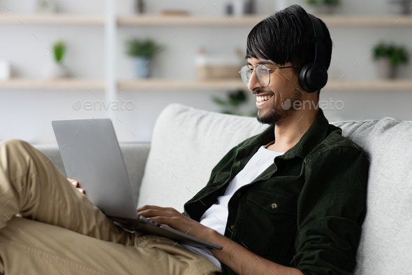 Positive indian guy chilling at home, using laptop and headset