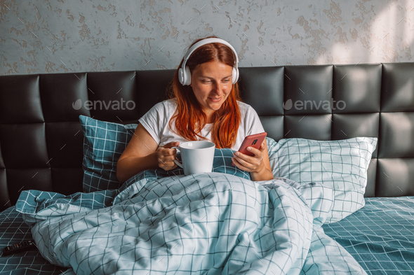 Woman wearing headphones listening to music enjoy new playlist rest relax sitting in bed