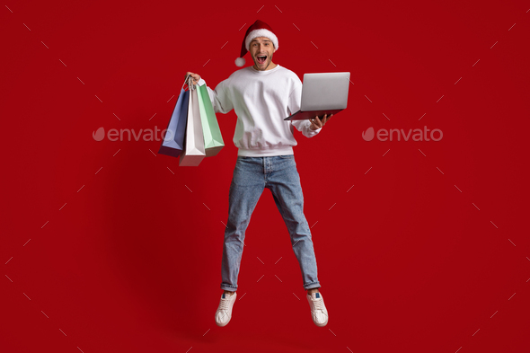 Online Sales. Excited Man Wearing Santa Hat Holding Laptop And Shopping Bags
