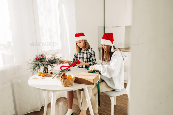 Two girls in Santa Claus hats are sitting at the kitchen table and packing sweet gifts to friends