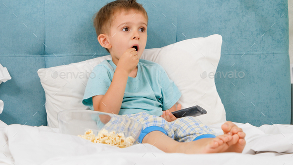 Little boy in pajamas eating popcorn in bed at morning and switching cartoons on TV with remote