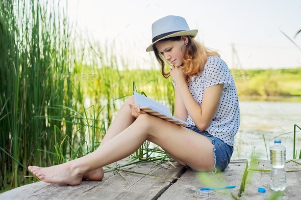 Teenage girl in hat reads diary sitting on shore of lake, nature landscape background