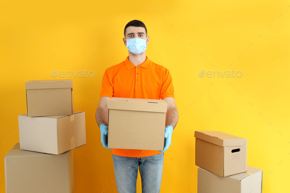 Delivery man in gloves and mask holds box on yellow background