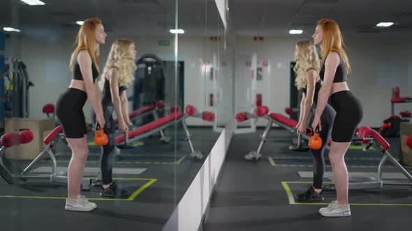 Side View of Confident Fit Cheerful Sportswomen Lifting Weights in Gym Reflecting in Mirror alt