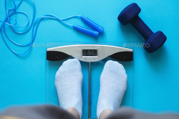Woman measures her weight on scale on a blue background with skipping rope and dumbbells