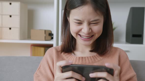 Excited young Asian woman playing games on smartphone in the living room at home. alt