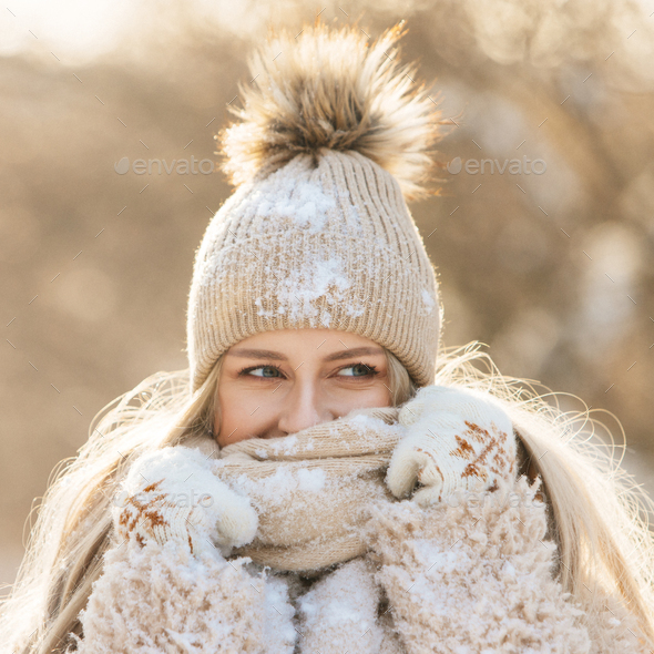 woman in beige hat with fur pompon, warm scarf, white gloves covered with snow at sunny winter day