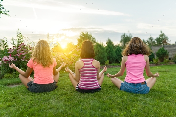 Group of young teenage girls practice yoga, meditate, sit in a lotus position on the grass near the