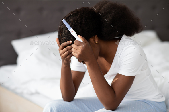 desperate african american lady sitting on bed with pregnancy test