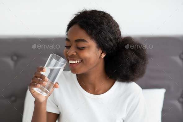 Happy young black woman drinking water in bed
