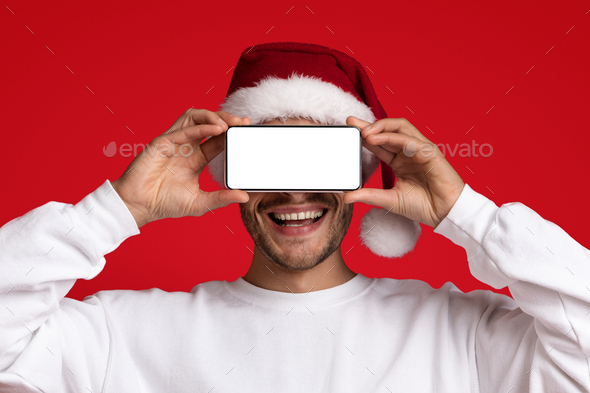 Online Ad. Smiling Man In Santa Hat Covering Eyes With Blank Smartphone