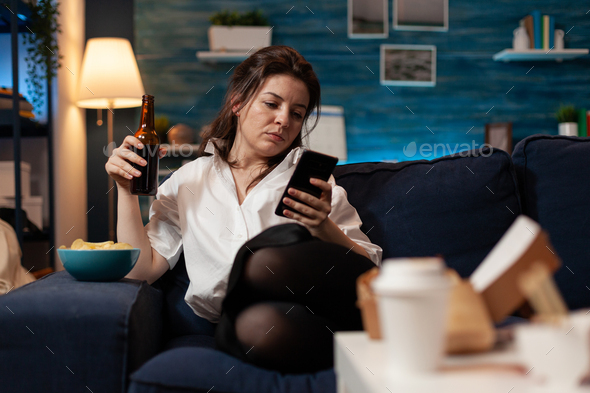 Woman relaxing on couch after work browsing social media on smartphone holding beer bottle