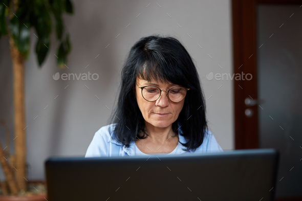 Mature woman writer or jiurnalist in glasses typing on laptop article