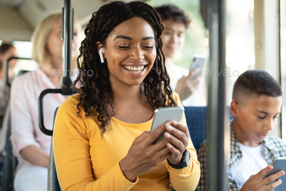 Happy black lady using smartphone wearing headphones in bus