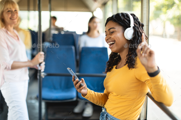 Excited black lady dancing in city bus wearing headphones