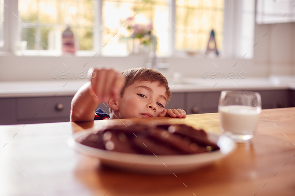Boy At Home In Kitchen Reaching Up To Take Cookie From Plate On Counter