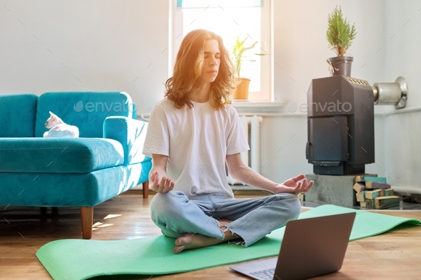 Guy teenager sitting in lotus position on yoga mat at home on floor with laptop