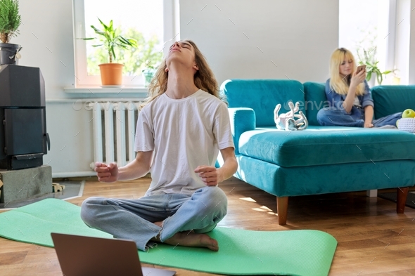 Guy teenager sitting in lotus position on yoga mat at home on floor with laptop