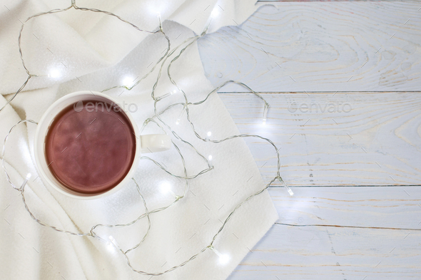 Cup of tea or coffee, knitted white blanket and garland on light wooden background.