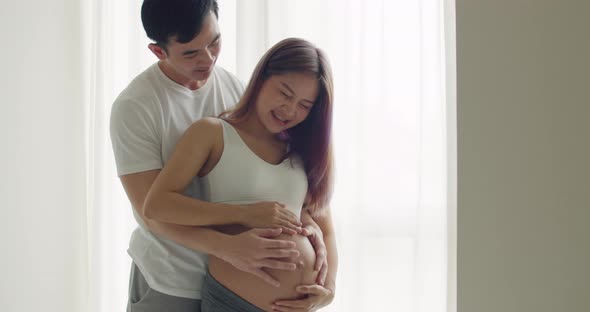 Asian husband touching his wife's pregnant belly while standing beside a window.