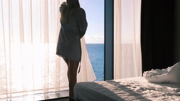 Blonde in a Gray Silk Bathrobe Opens Curtains on the Window View of Sea and the Sun
