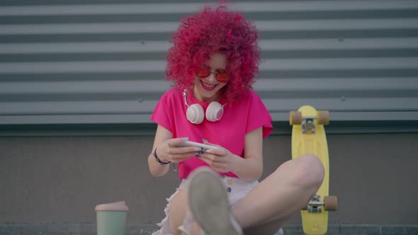A Pink Curly Teenager is Playing Mobile Game on Smartphone Outdoors alt