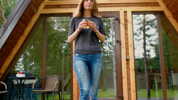 Woman Comes Out On The Porch Of A Small Wooden House With A Glass Of Juice