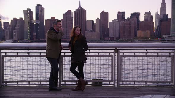 Couple in New York City stand by river talking with skyline in background