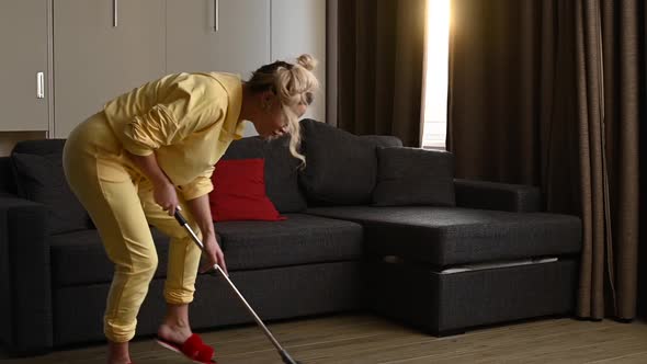 Happy Blonde Woman in a Yellow Hoodie with a Mop Cleaning the House and Dancing alt