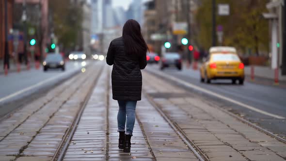 a Brunette with Long Hair in a Knitted Sweater and a Dark Coat Walks Along the Tram Tracks on a alt