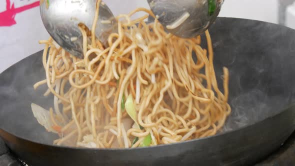 A Female Cook's Hand Frying a Plate of Thai Noodles in a Frying Pan at an Outdoor Food Festival alt