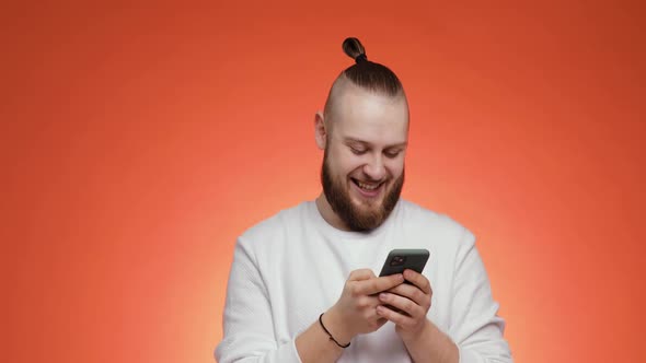 Bearded Man with Hair in Tail Laughing Typing on Smartphone on Red Background