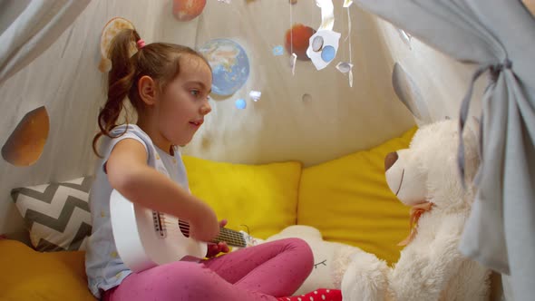 Little Girl Plays a Toy Guitar for a Teddy Bear Sitting in Tent Home alt