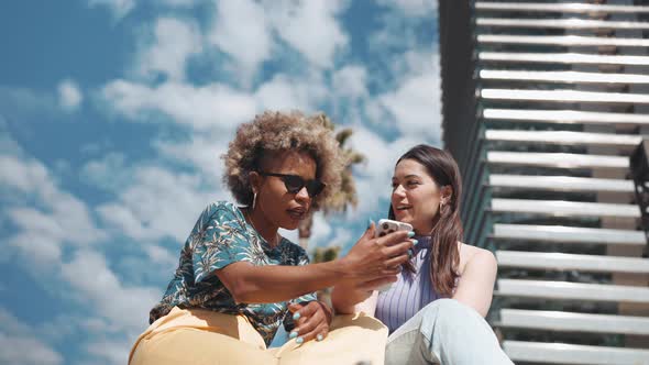 Surprised sunny multiethnic women talking and looking at phone outdoors alt