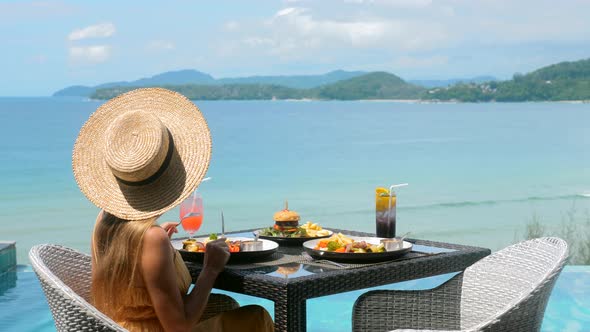 Woman Eating Dinner Sitting at Restaurant Terrace Near Pool with Blue Sea View alt