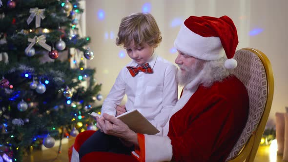 Side View Santa Reading Book for Little Cute Caucasian Boy Indoors in Living Room alt