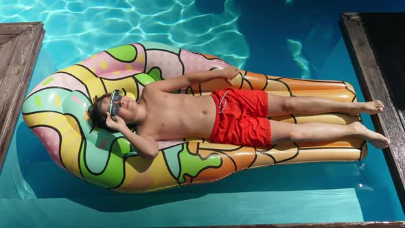 Top View of Relaxed Little Boy in Sunglasses Lying on Swimming Mattress Sunbathing in Swimming Pool alt